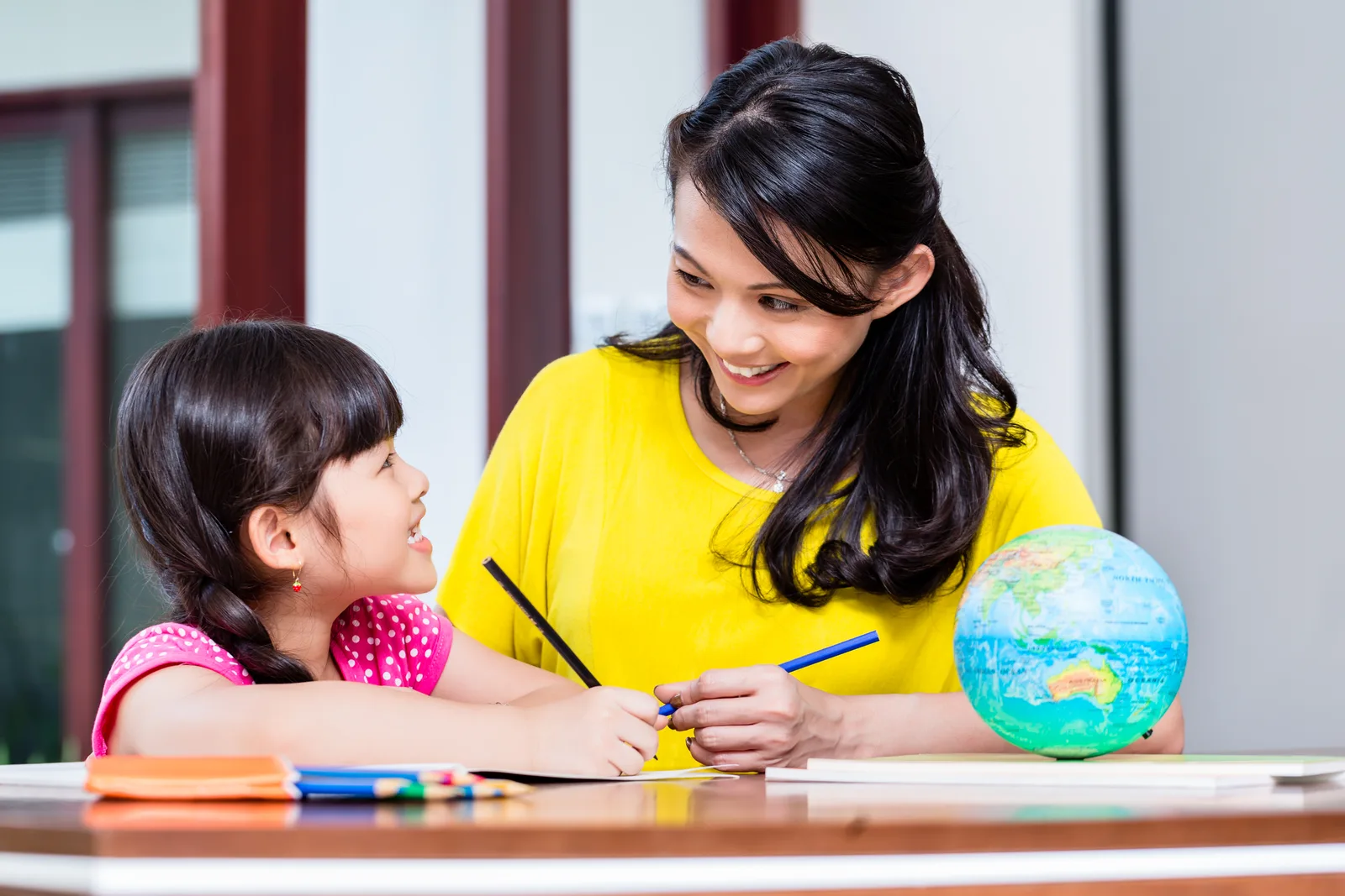 A mom and her young daughter smiling at each other over schoolwork, a globe on the table